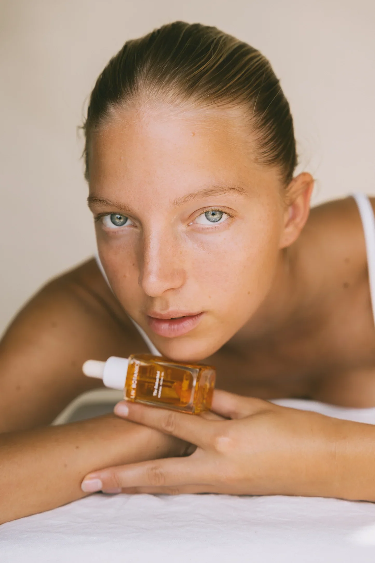 Close-up of a woman with blue eyes and light brown hair in a sleek ponytail, holding a small bottle of skincare product, lying on a white surface with a neutral background.