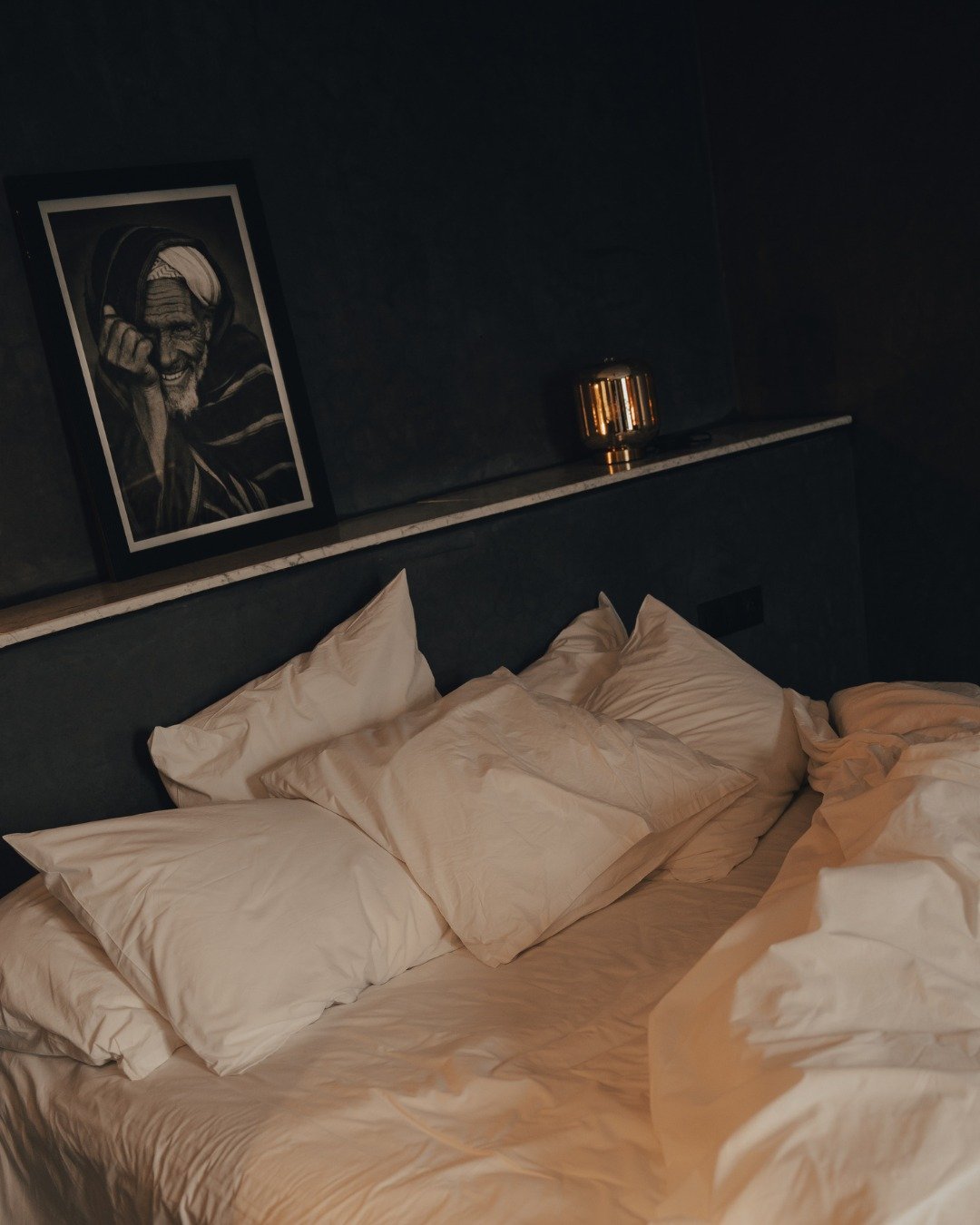 A messy bed with white sheets and multiple pillows, a black headboard, and a dark wall with a framed black-and-white portrait of an elderly man wearing headphones. There is a small gold-colored lamp on the bedside shelf.