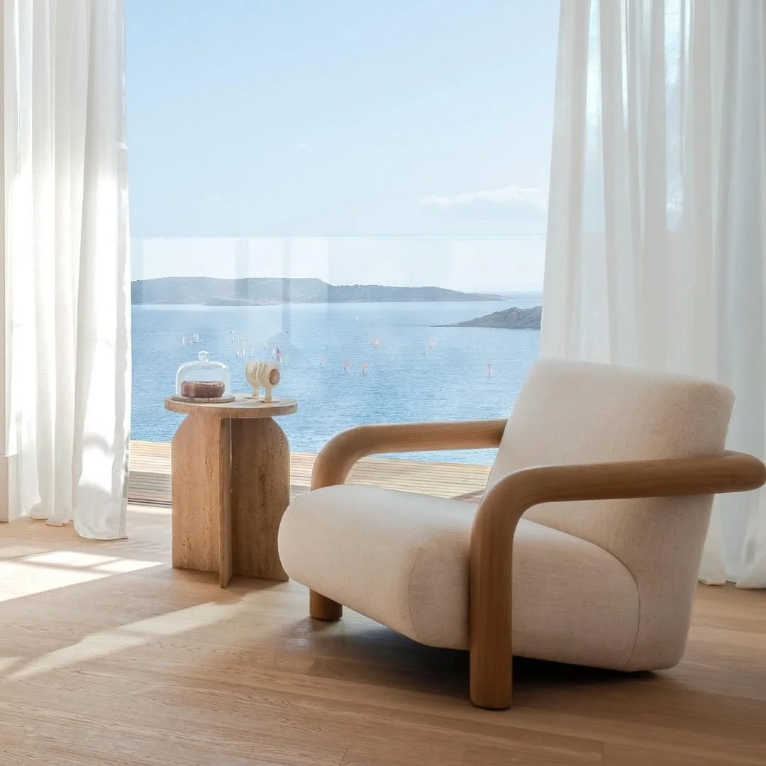 A cozy armchair with wooden armrests set by large glass doors opening to a view of a lake or sea with small sailboats and distant islands, with white curtains framing the scene.