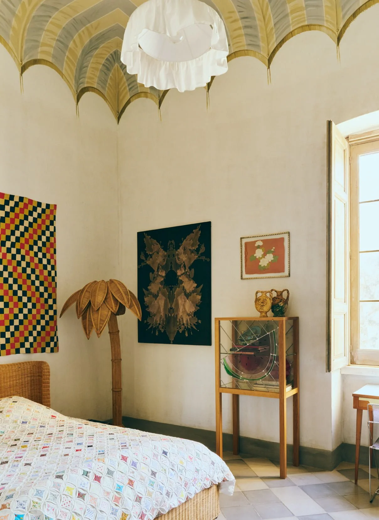 A bedroom corner featuring a bed with a colorful quilt, a bamboo leaf-shaped lamp, a dark butterfly-themed artwork, a small wooden display case with decorative items, and framed floral artwork on a cream wall. There's an open window with wooden shutters letting in natural light.