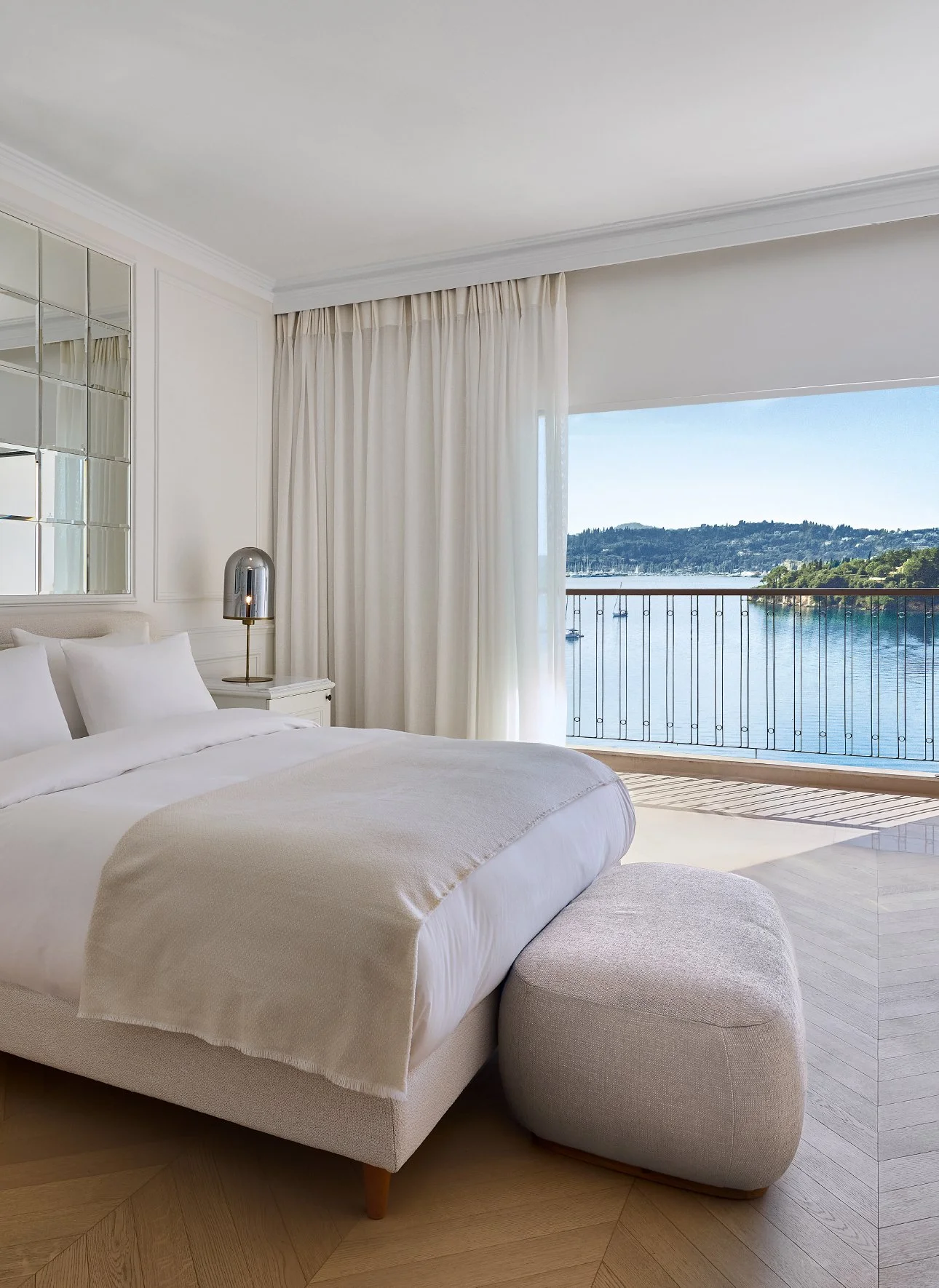 Bedroom with large bed, white bedding, round ottoman, nightstand with metallic lamp, large window with beige curtains, overlooking a water view with boats and hills.