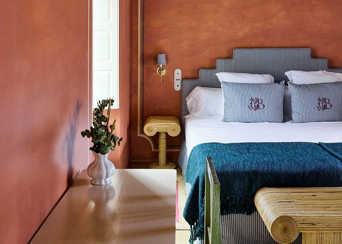 A bedroom with pinkish-orange walls, a striped headboard, white and striped pillows, a white bedside table with a vase and green foliage, a rattan side table, and a bed with a teal blanket.