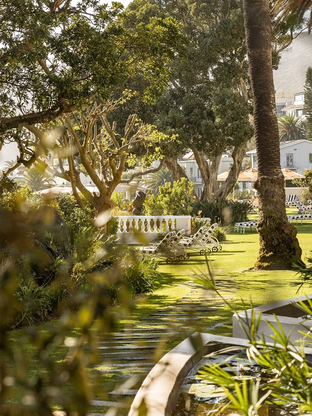 A lush outdoor garden area with green grass, trees, and lounge chairs, with a pathway leading through the garden.