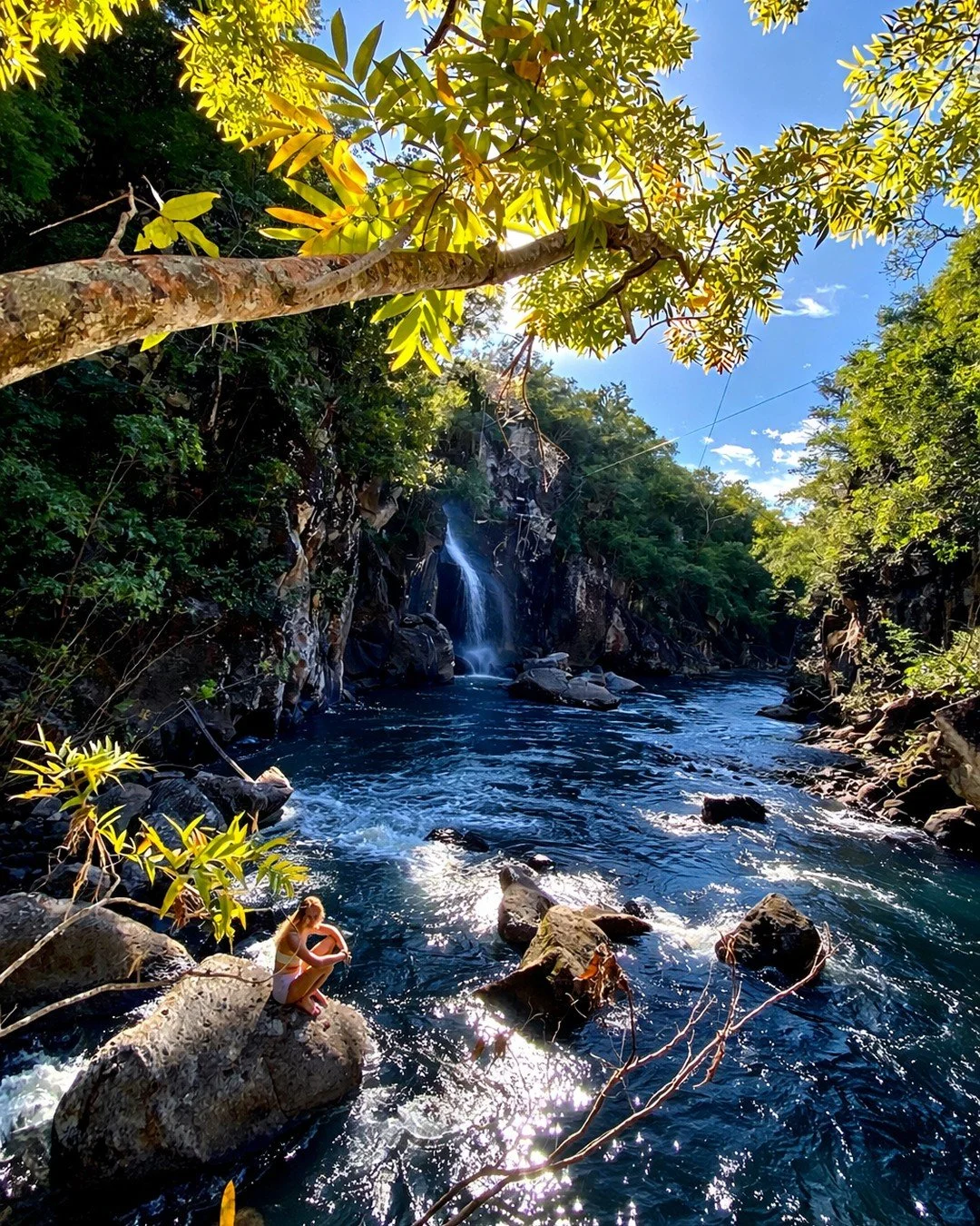 A woman sitting on a large rock by a flowing river surrounded by green trees, a small waterfall in the background, and bright blue sky overhead.