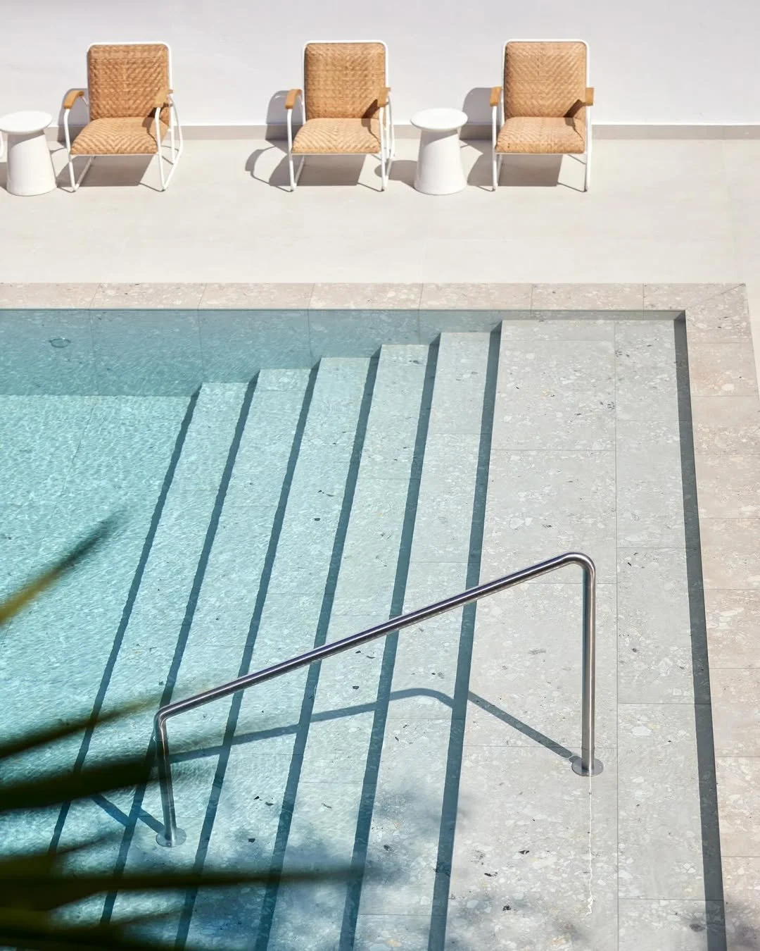 Empty swimming pool with steps and a metal handrail, surrounded by a white tiled deck and three wicker chairs with beige cushions and small white side tables.
