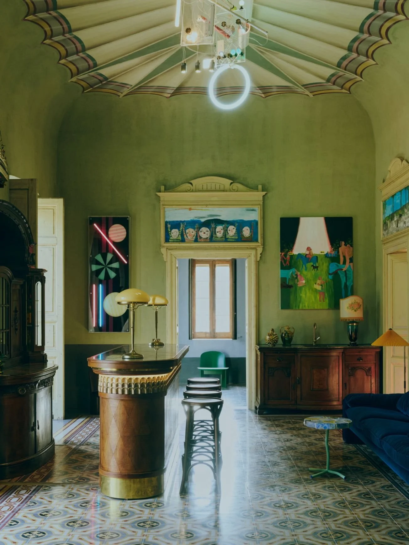 Interior of a stylish, artistically decorated room with patterned tile floor, green walls, and eclectic art pieces including a colorful painting and a neon light installation. A wooden bar counter and various vintage furniture are present, with a window at the back letting in natural light.