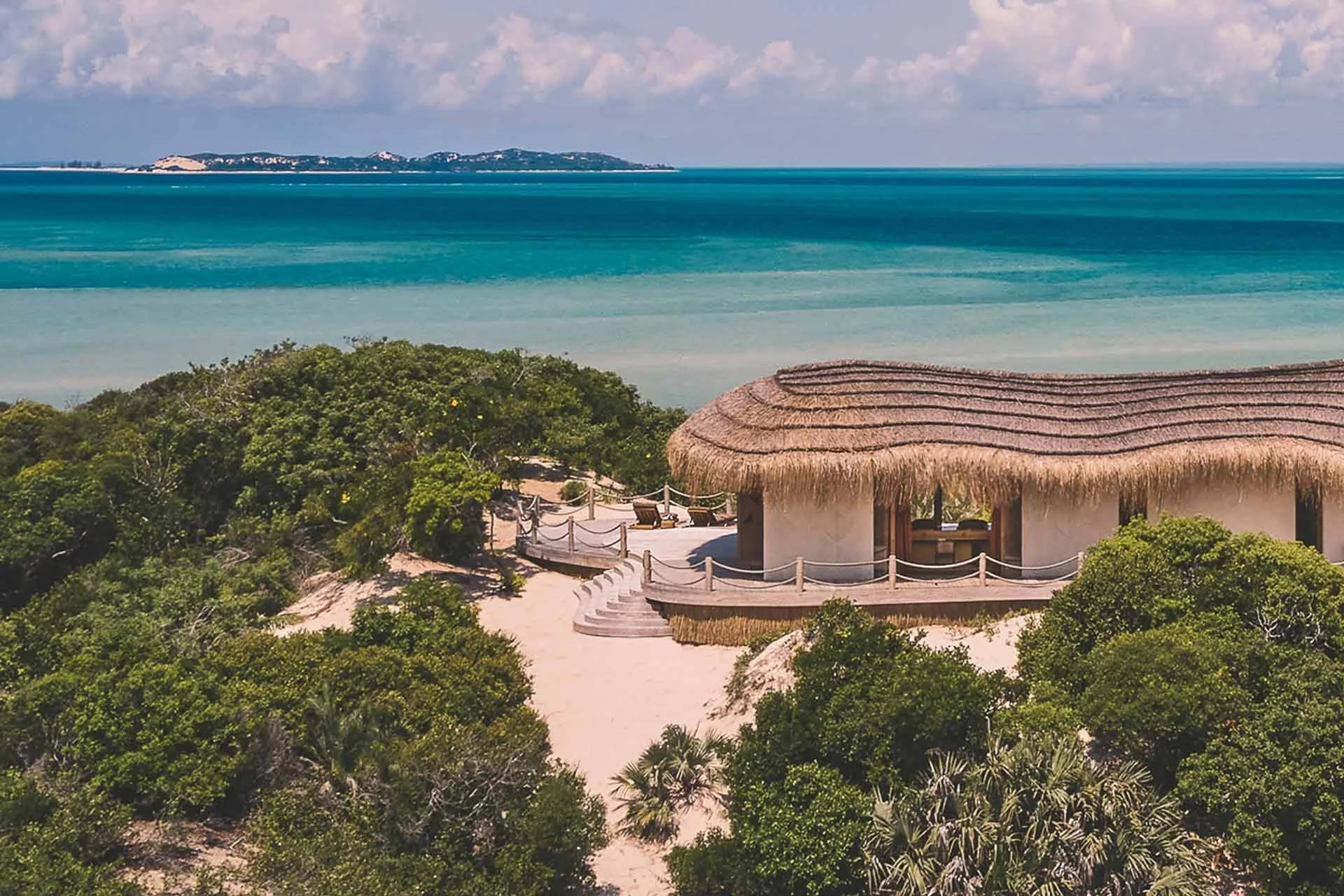 A beach resort with a thatched-roof hut surrounded by lush green trees, overlooking a large body of turquoise water with a distant landmass under partly cloudy skies.