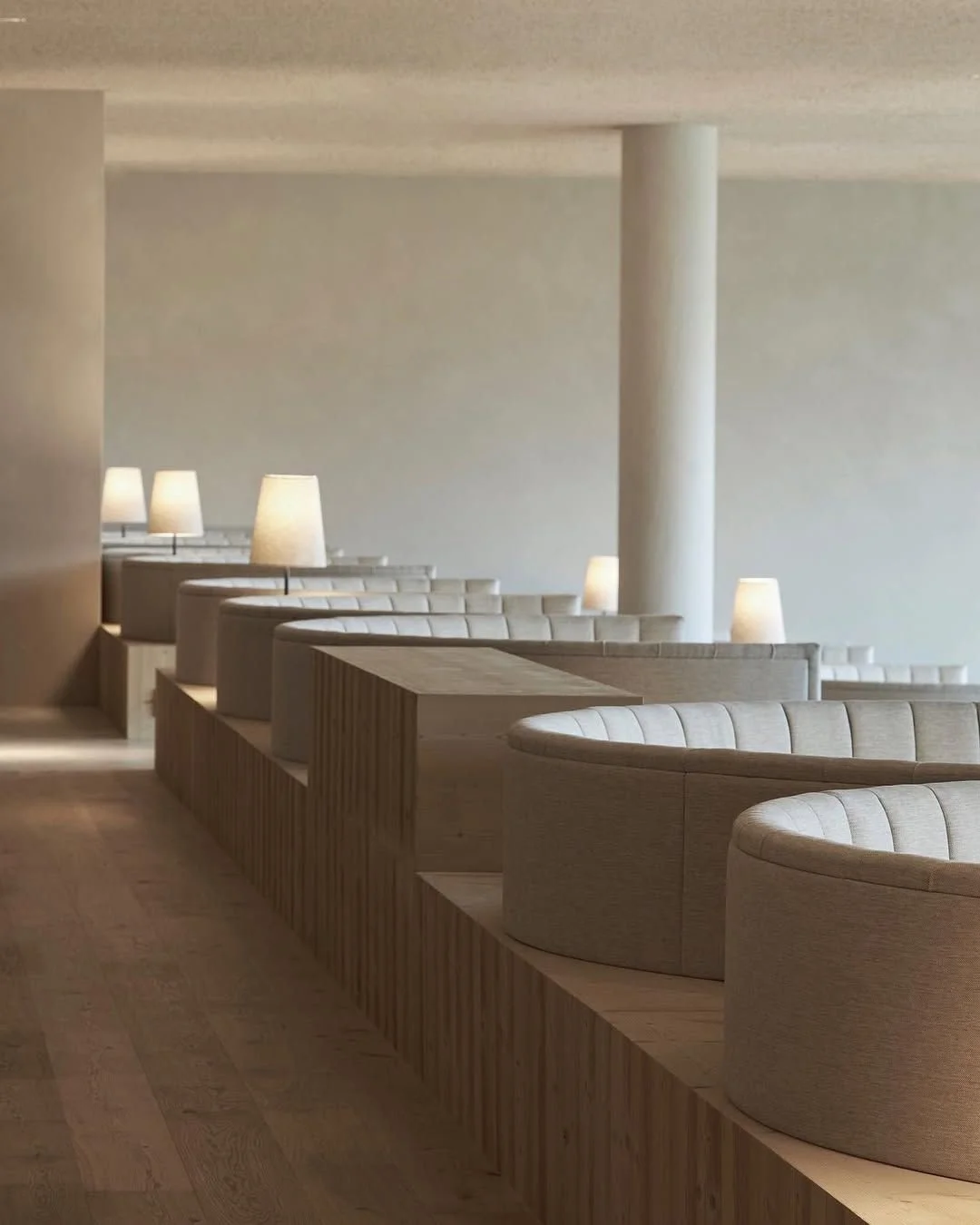 A modern, minimalist interior with curved beige booths, wooden accents, and small lamps with white shades on tables.