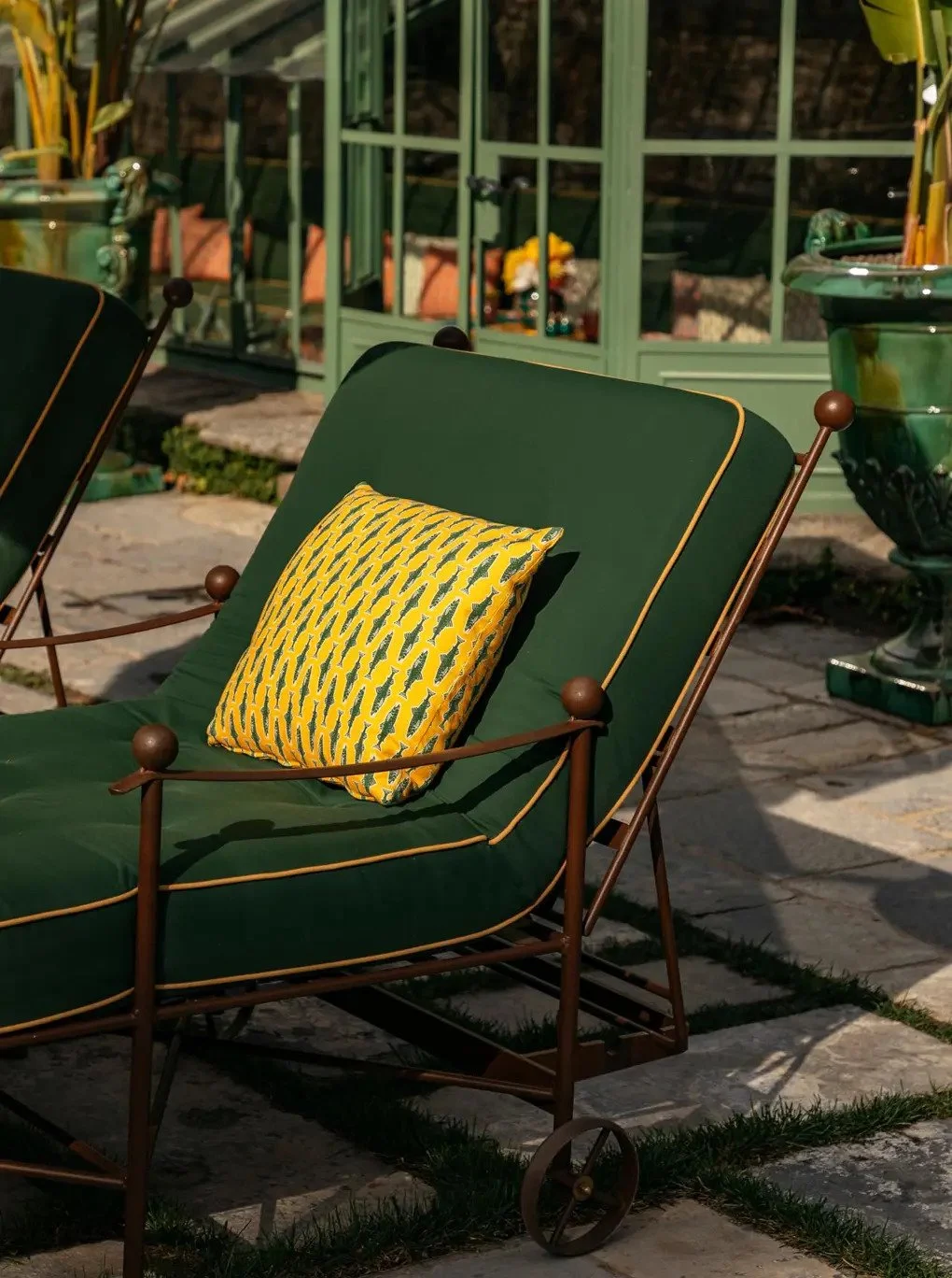 A green outdoor lounge chair with a yellow patterned pillow, situated on a stone patio in front of a green glass-paneled structure.