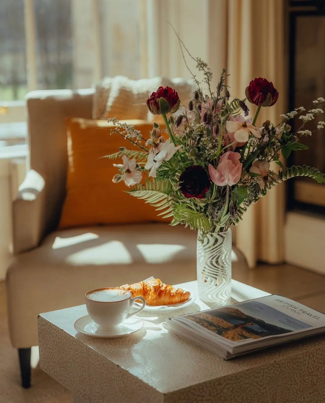 A cozy living room scene with a side table holding a vase of mixed flowers, a cup of coffee, a croissant, and a travel magazine. A cushioned armchair with a beige throw pillow is in the background, next to a window with sunlight streaming in.