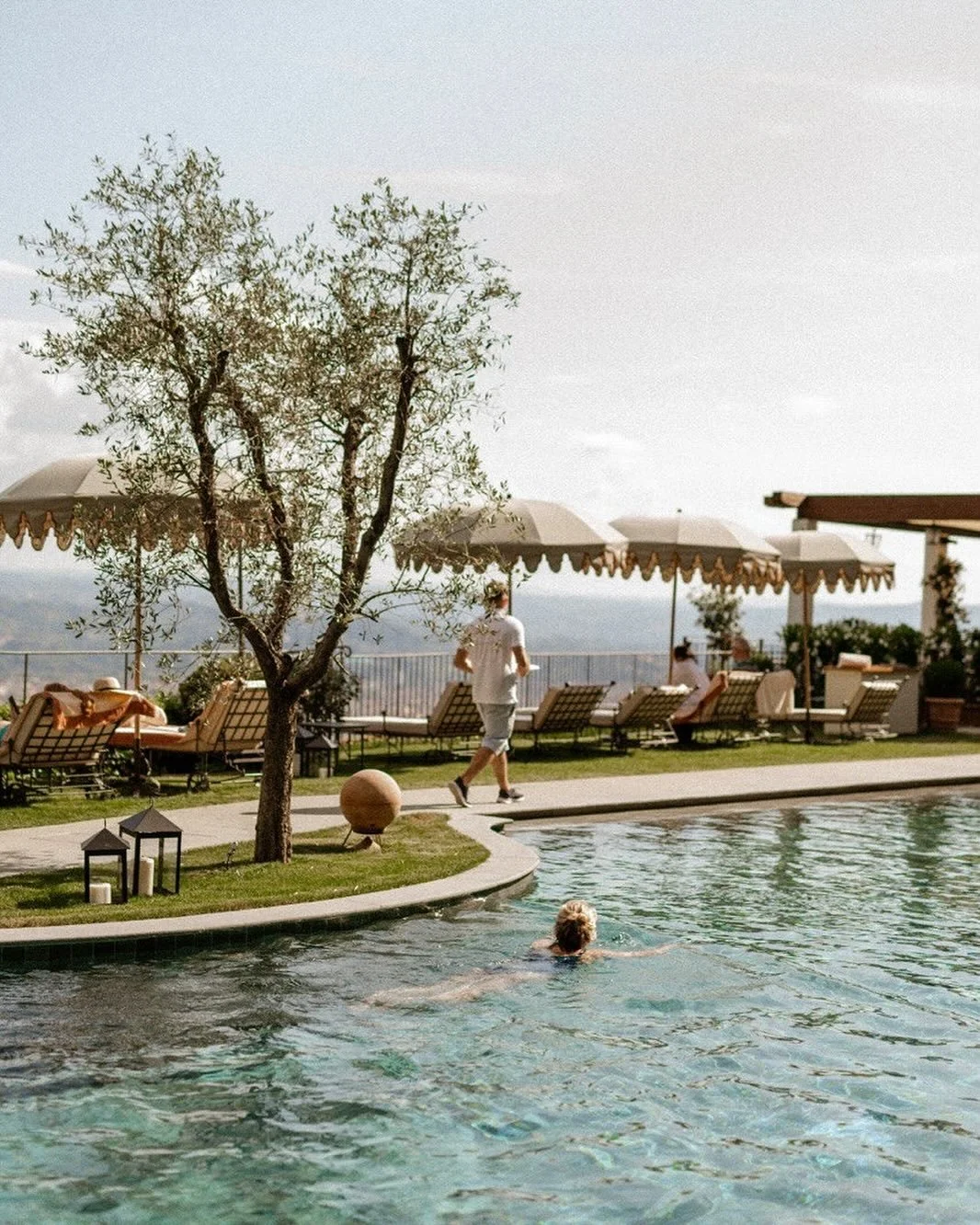 A swimming pool with a person swimming, garden chairs with umbrellas, and a woman walking along the poolside. Trees and a distant view in the background.