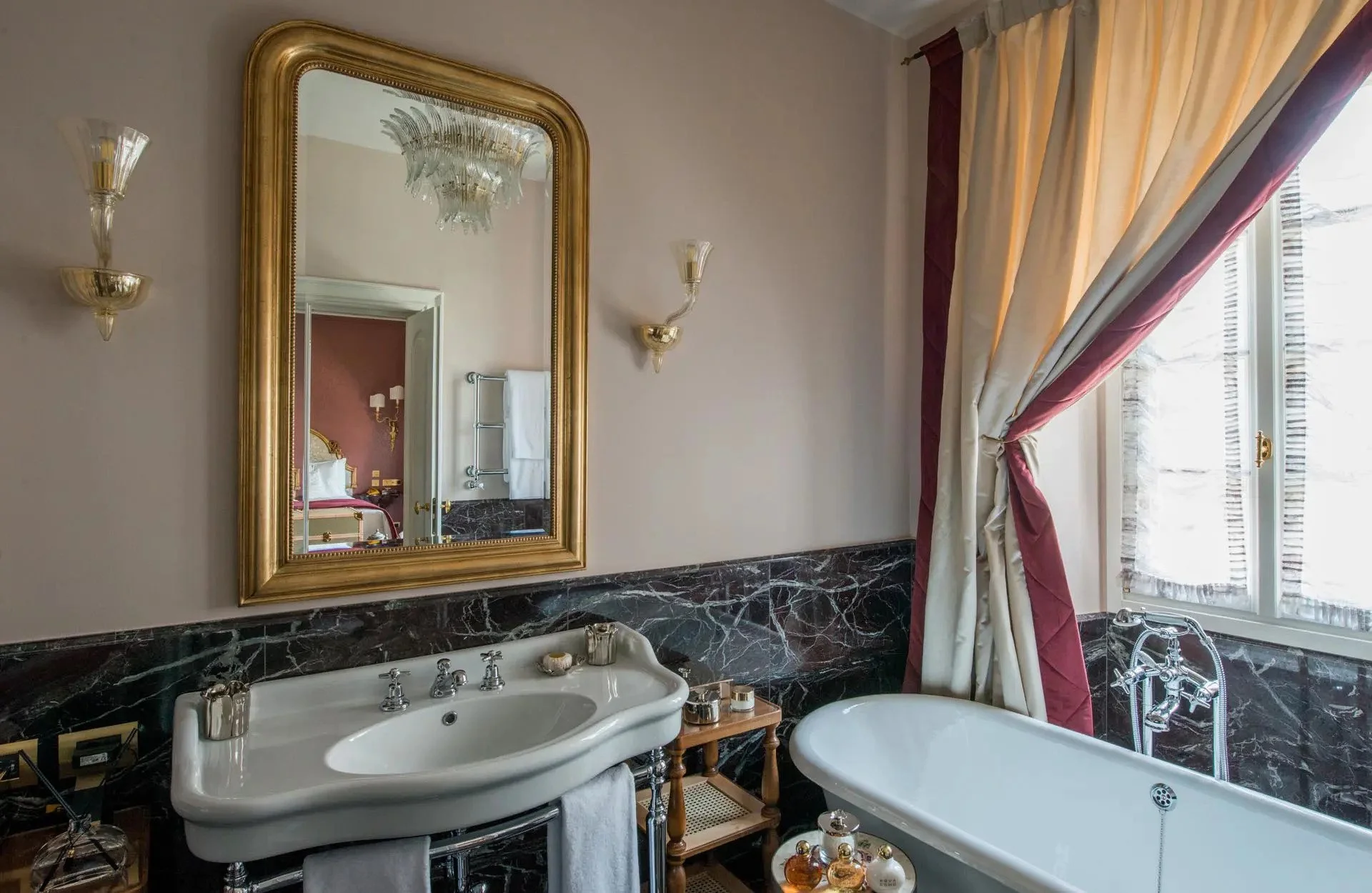 Luxurious bathroom with a gold-framed mirror, marble sink, and clawfoot bathtub near a window with red and cream curtains.