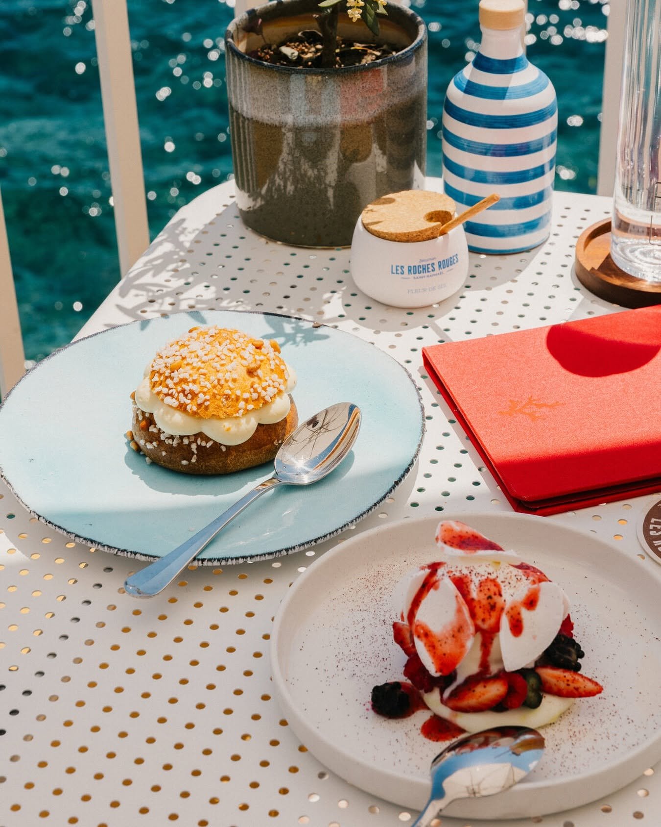 A table with a dessert topped with whipped cream, strawberries, and syrup in a white bowl, along with other desserts and drinks, set outdoors near water. There is also a small jar labeled 'Les Roches Rouges' and a striped vase, a red book or menu, and beverages.