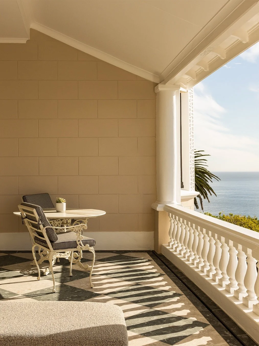 A balcony with a round table and two chairs, overlooking the ocean and sky, with palm trees in the background.