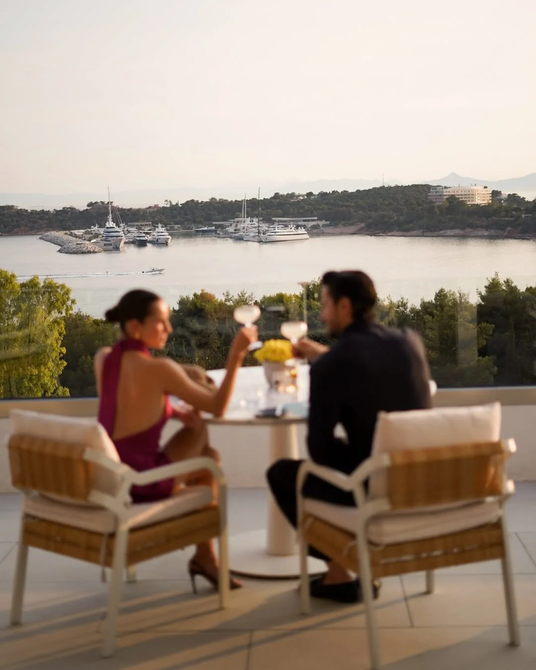 A couple dressed formally, toasting drinks at a table on a balcony overlooking a harbor with yachts and boats, and green trees and buildings in the background.