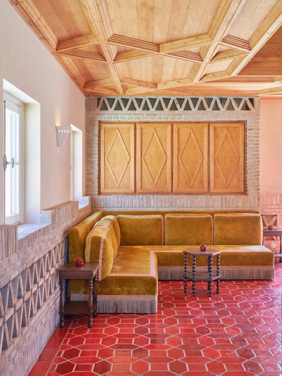 Interior room with a wooden coffered ceiling, brick and plaster walls, a built-in wooden panel, mustard-colored sectional sofa, small wooden side tables, and windows with white framing, red hexagonal tile flooring.