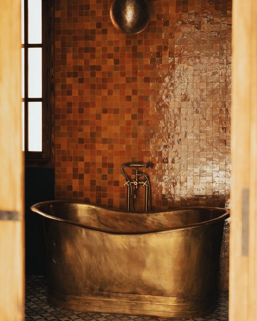 A vintage-style copper bathtub with a curved shape, set against a brown tiled wall with small square tiles, in a bathroom with a mosaic tile floor, and a window with wooden framing on the left.