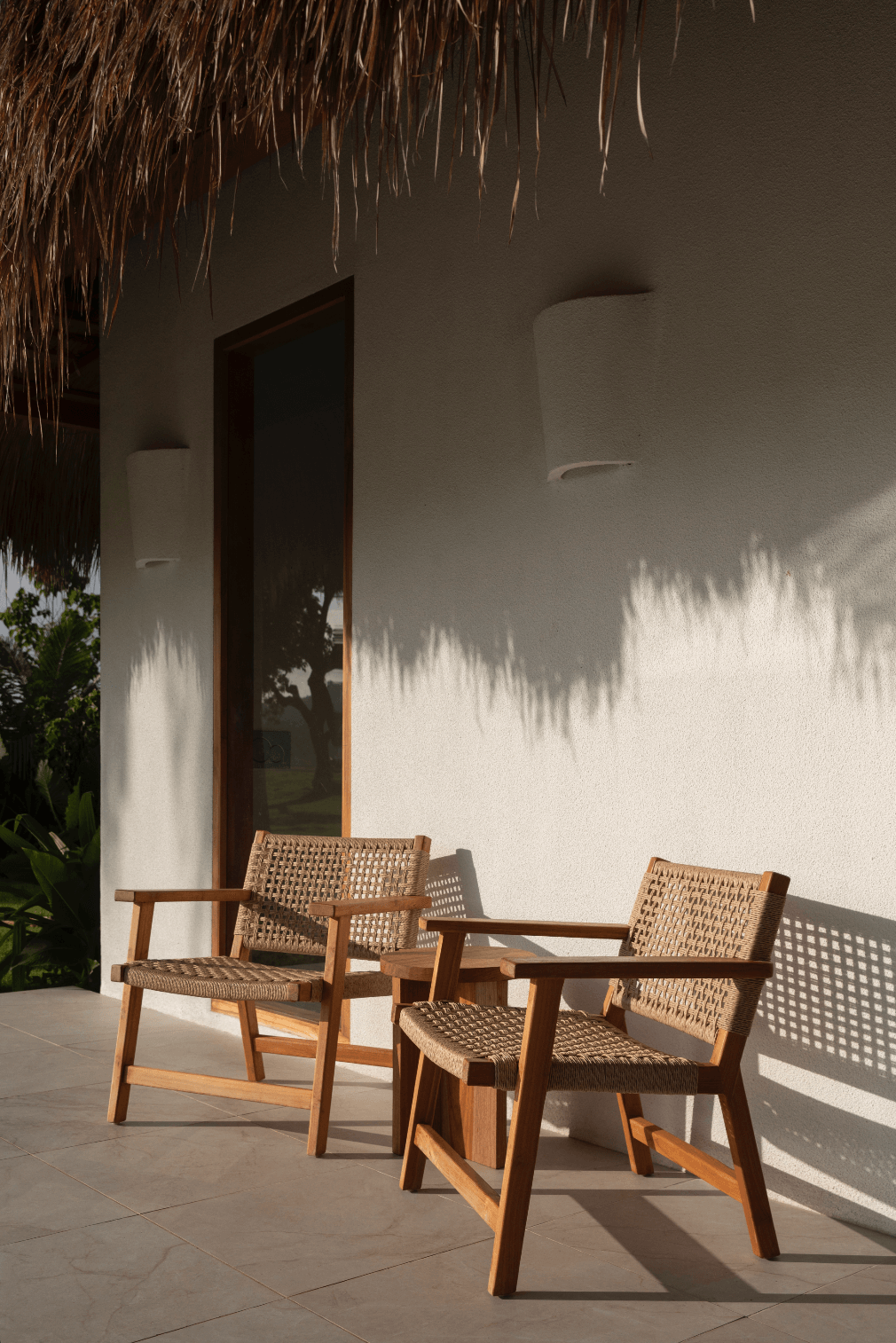 Two woven wooden chairs with armrests on a tile patio, shadowed by sunlight, against a white wall with a window and thatched roof overhang.