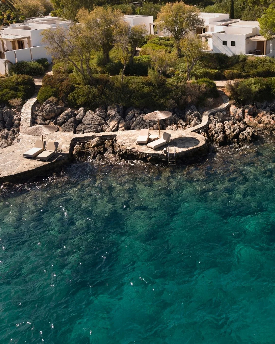 Beachside seating area with lounge chairs and umbrellas on rocky platform near clear blue water, surrounded by lush green trees and modern white buildings in the background.