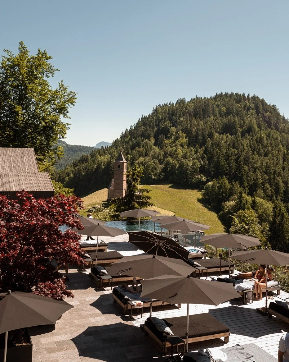 Outdoor pool area with lounge chairs and umbrellas overlooking a green hillside with a church tower and forested mountains in the background.