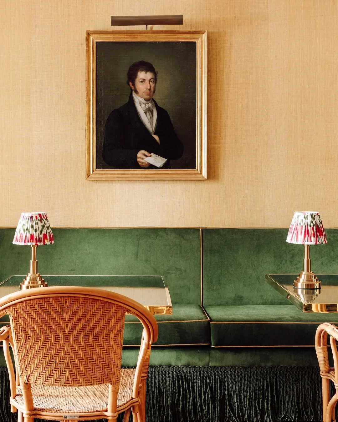A painting of a man in historical attire hanging on a beige wall in a room with green seating, a wicker chair, and two lamps with red and green patterned shades.