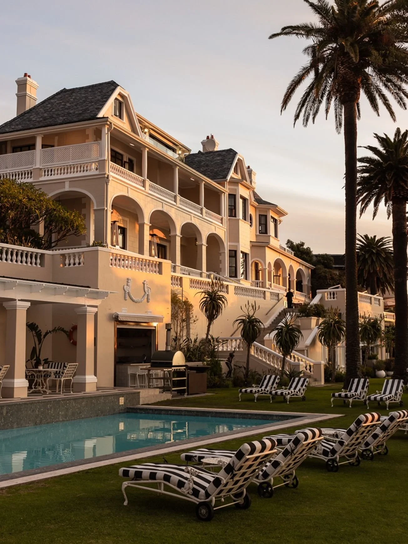 Elegant multi-story house with white exterior, arches, and large balconies. It has a swimming pool in the backyard, surrounded by lounge chairs and palm trees, during golden hour.