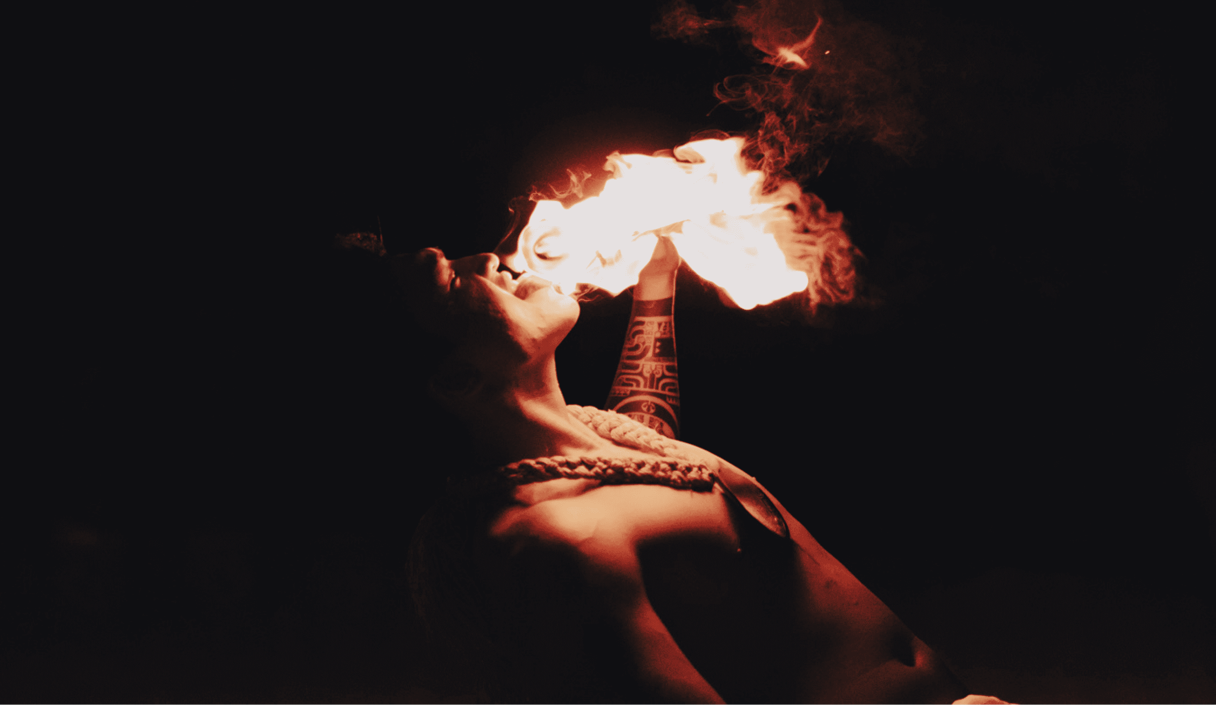 A person with tattoos on one arm, holding a lit torch in their mouth, creating a large flame and smoke, against a dark background.