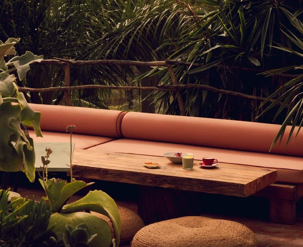 Outdoor patio with a long wooden table, pink cushioned seating, and bowls and cups on the table, surrounded by lush green tropical plants.