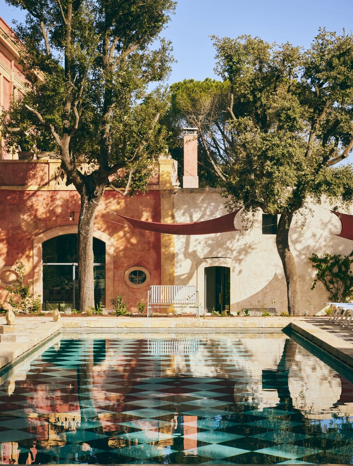 A courtyard with two large trees, colorful house walls, a swimming pool with a black and white patterned floor, and shaded fabric canopies.