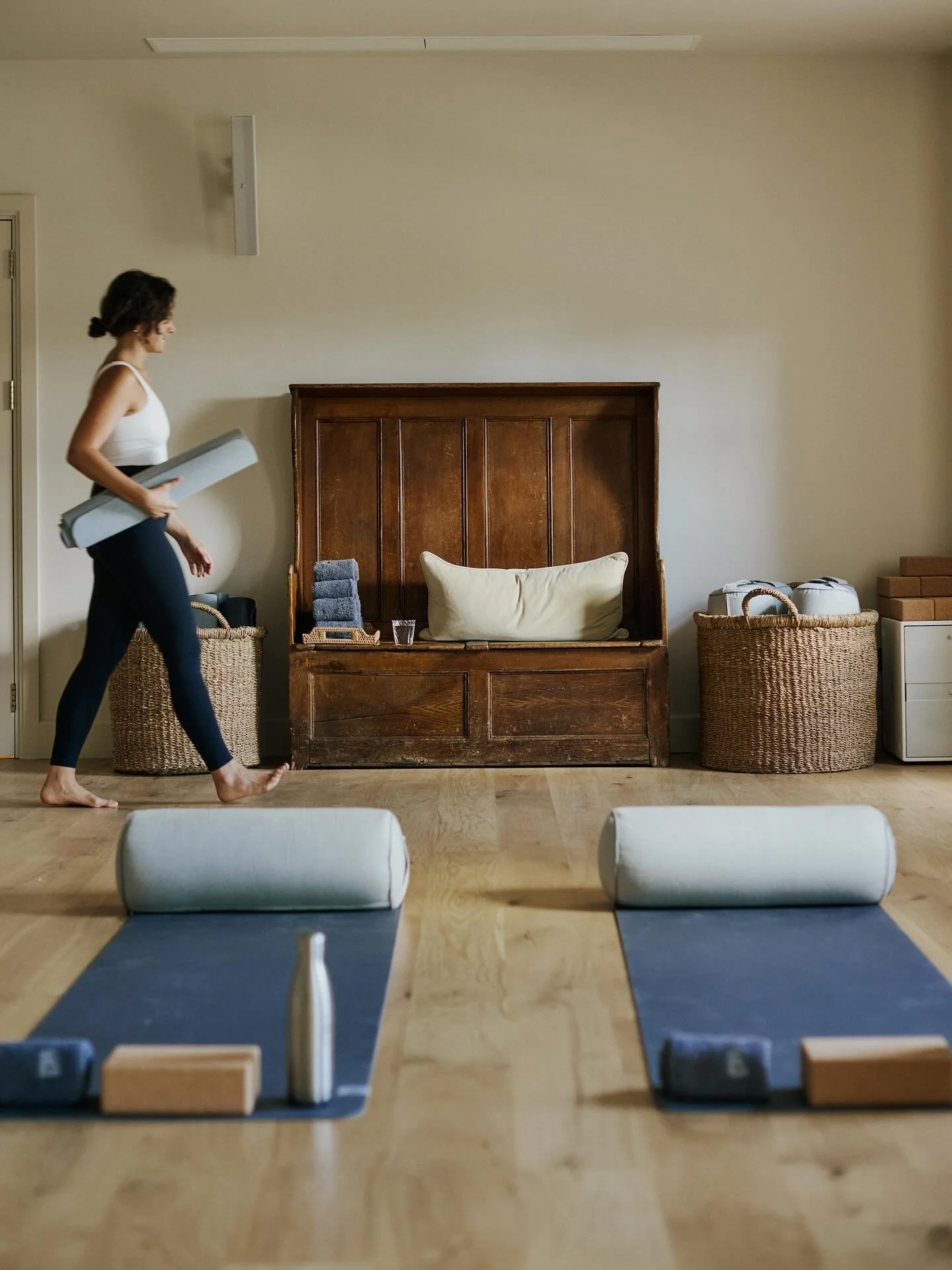 A woman in black leggings and a white tank top walking past yoga mats and blocks in a room with wooden floors and neutral-colored walls.