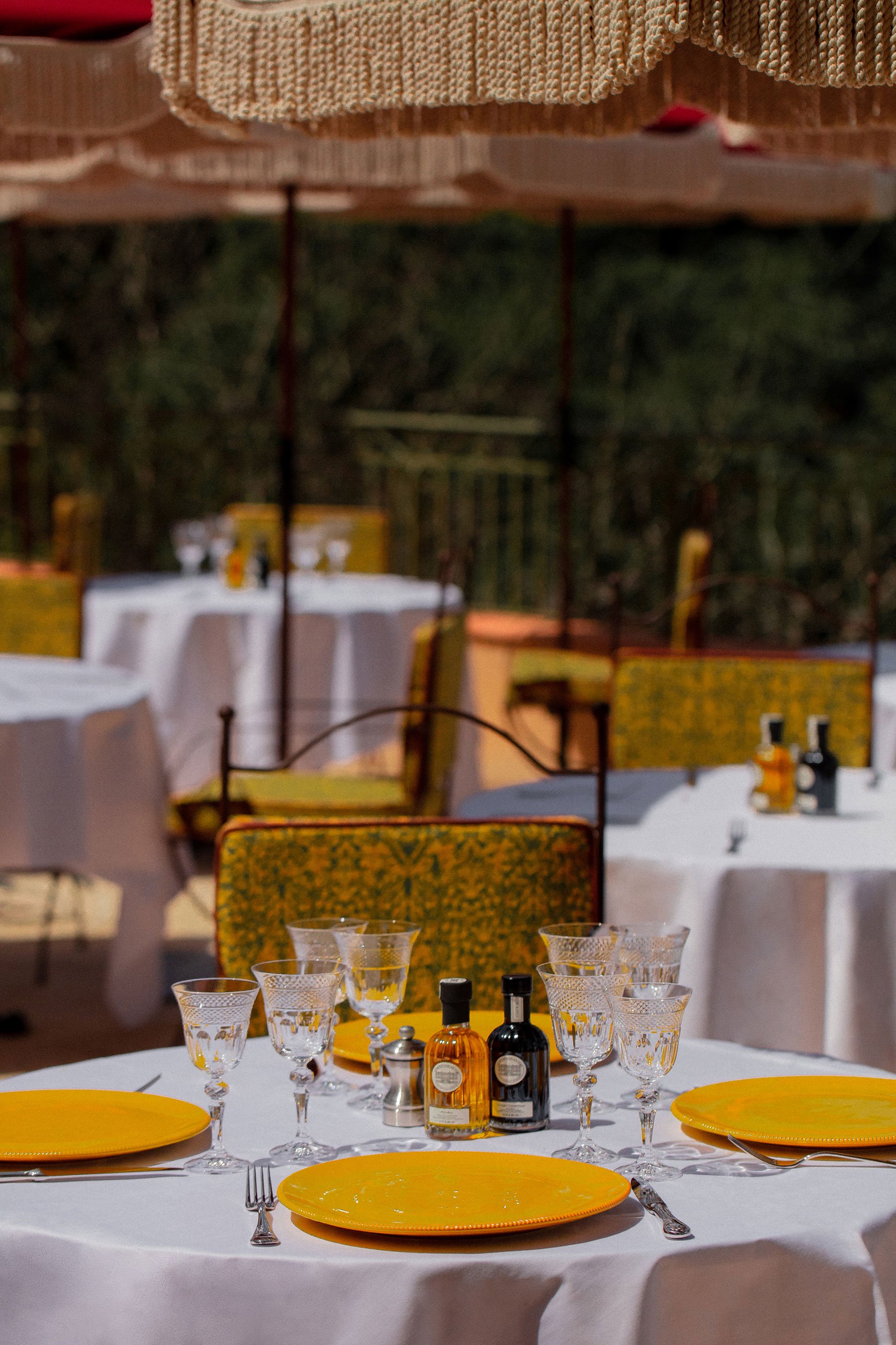 A round banquet table set for a meal with yellow plates, crystal glasses, and silverware. The table has salt, pepper, and bottles of sauces or oils. In the background, there are white tablecloths and yellow chairs, with an outdoor setting that includes trees and umbrellas.