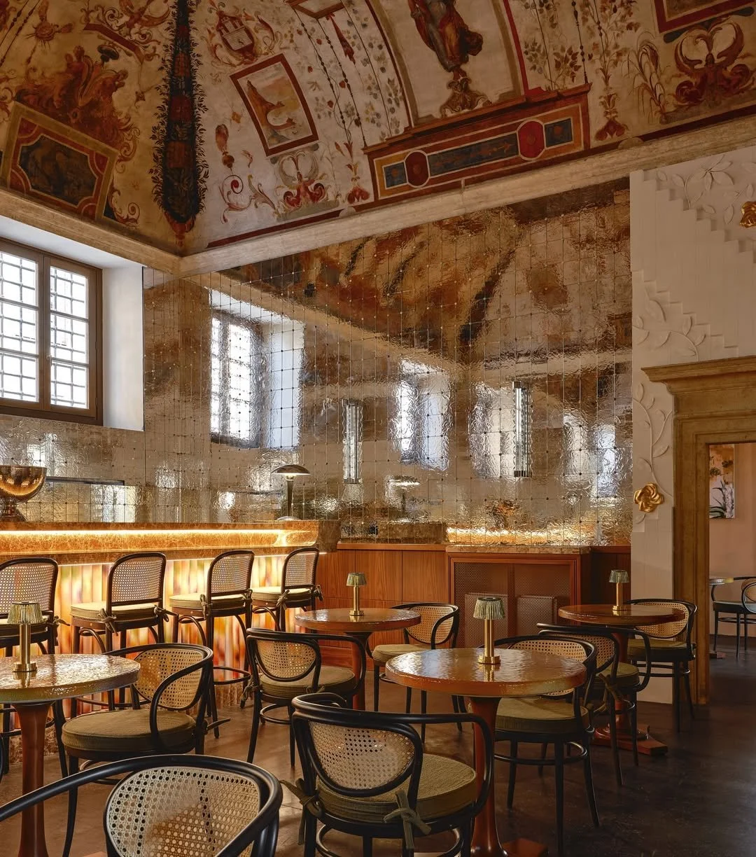 Elegant dining area with wooden tables and rattan chairs, gold lamps on tables, and a gold mirrored wall. The ceiling features ornate, colorful paintings and decorative motifs.