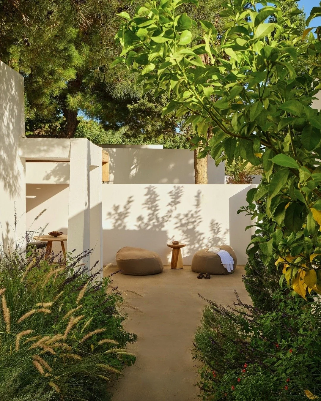 A peaceful outdoor patio with beige bean bag chairs, a wooden side table, and lush green trees and bushes surrounding a white wall.