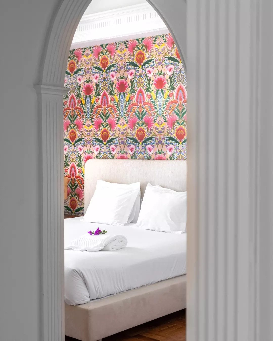 View of a neatly made bed with white linens, two pillows, and a towel with a small purple flower on top. The bed is viewed through an archway with white, decorative molding. Behind the bed, there is vibrant pink, purple, yellow, green, and orange floral wallpaper.
