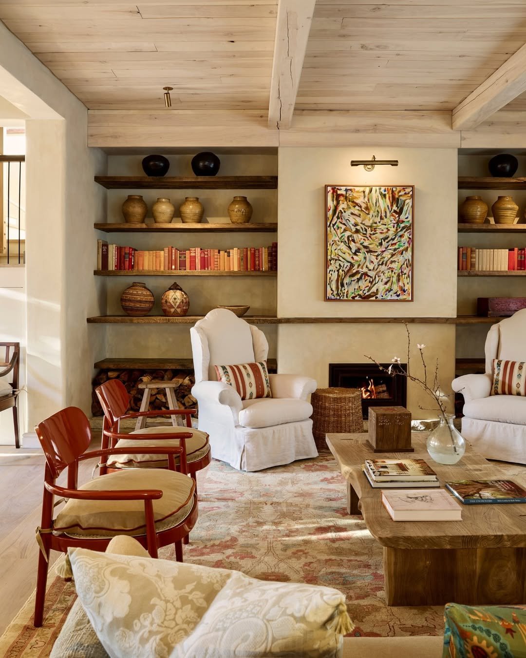 Living room with white armchairs, red wooden chairs, a wooden coffee table, a fireplace, and built-in shelves with books and decorative vases, wooden ceiling with beams and an abstract painting on the wall.