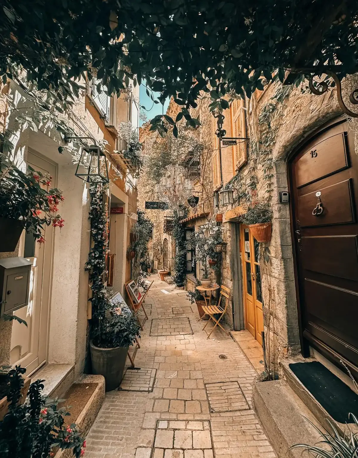 A narrow cobblestone alley lined with outdoor cafe seating, potted plants, and decorative lighting. Ivy and flowers climb the walls of stone and stucco buildings, creating a charming, European ambiance.