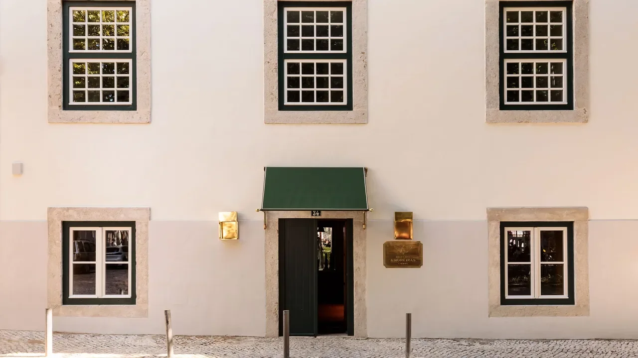 Front view of a white building with six windows framed in stone, three on the upper floor and three on the lower floor. The building has a green awning over the entrance door, and two gold-colored wall-mounted lamps beside it. There is a gold plaque next to the door.