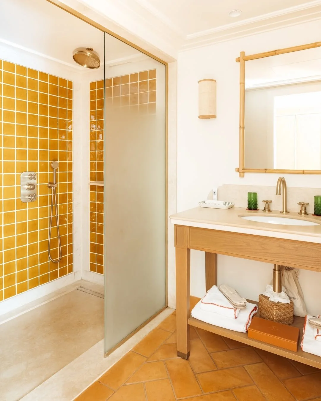 A bathroom with a walk-in shower with orange tiles, and a vanity with a mirror, green cups, towels, and toiletries.