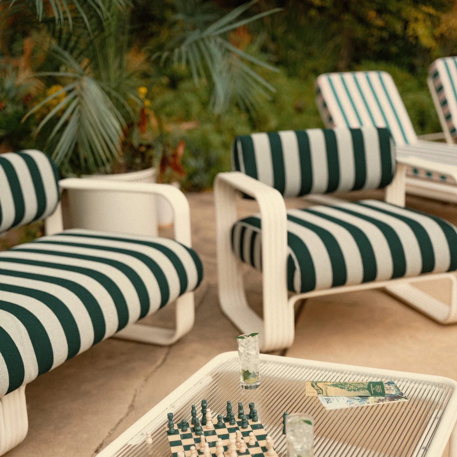 Outdoor patio area with white furniture featuring green and white striped cushions. A table in the foreground displays a chessboard with chess pieces and a glass of drink. In the background, lush green plants and trees are visible.