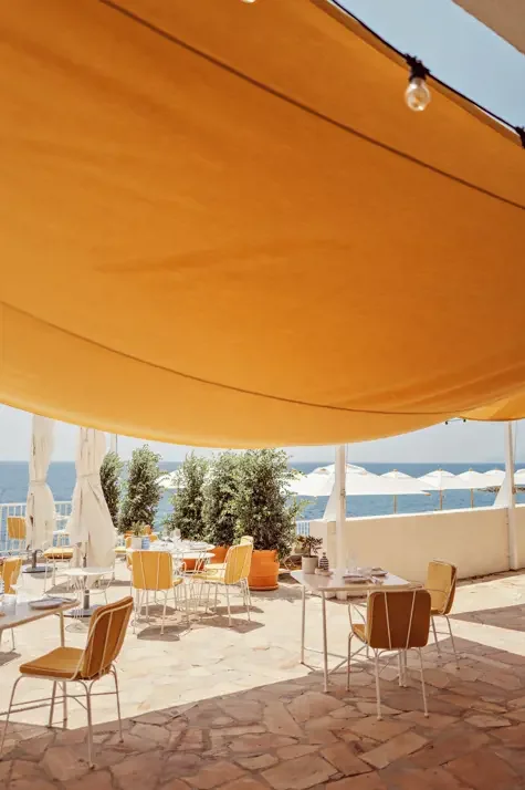 Outdoor patio dining area with tables and chairs, large canopy overhead, view of the ocean with umbrellas in the background, and potted plants.