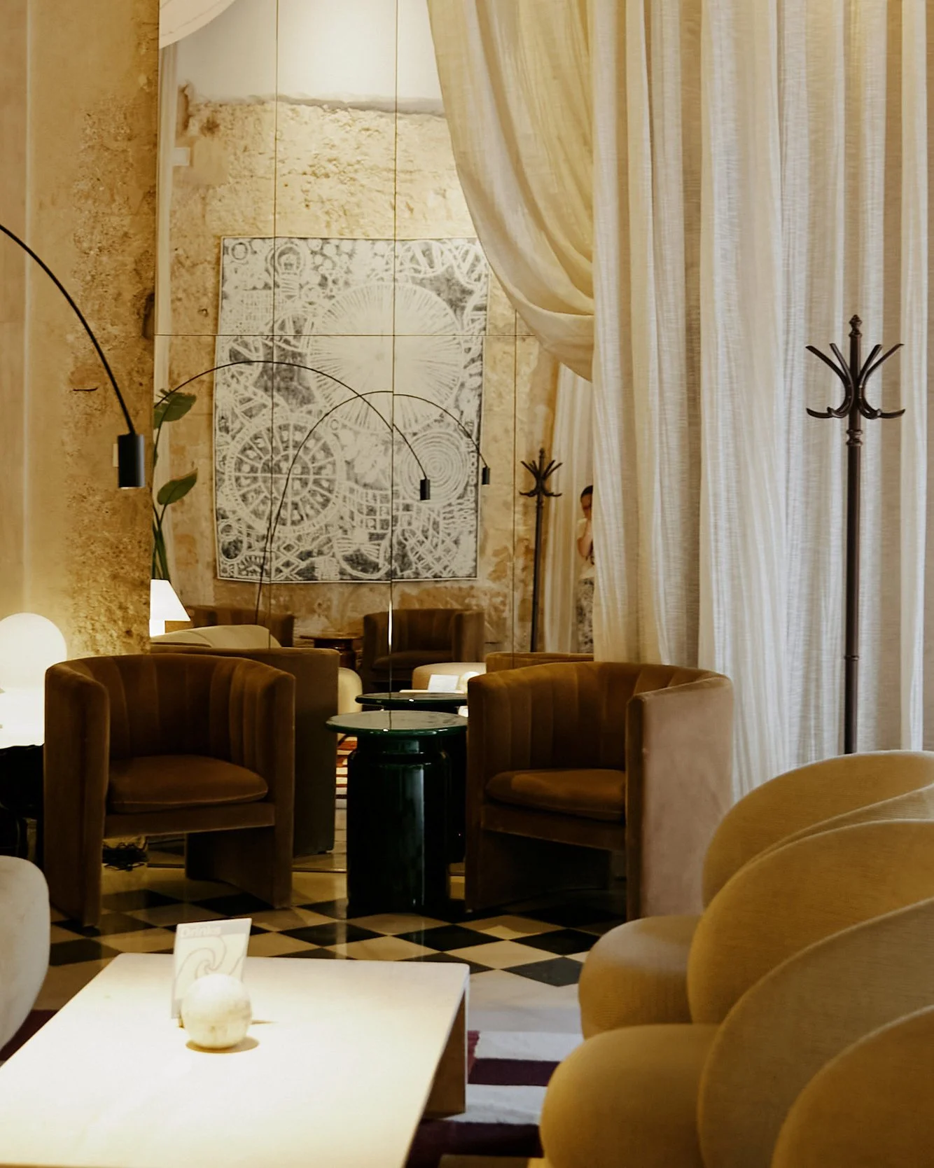 A cozy, stylish living room or lounge with beige and brown furniture, a white coffee table with a decorative ball, large curtains, and warm lighting. There is a textured wall with artwork and a mirror.