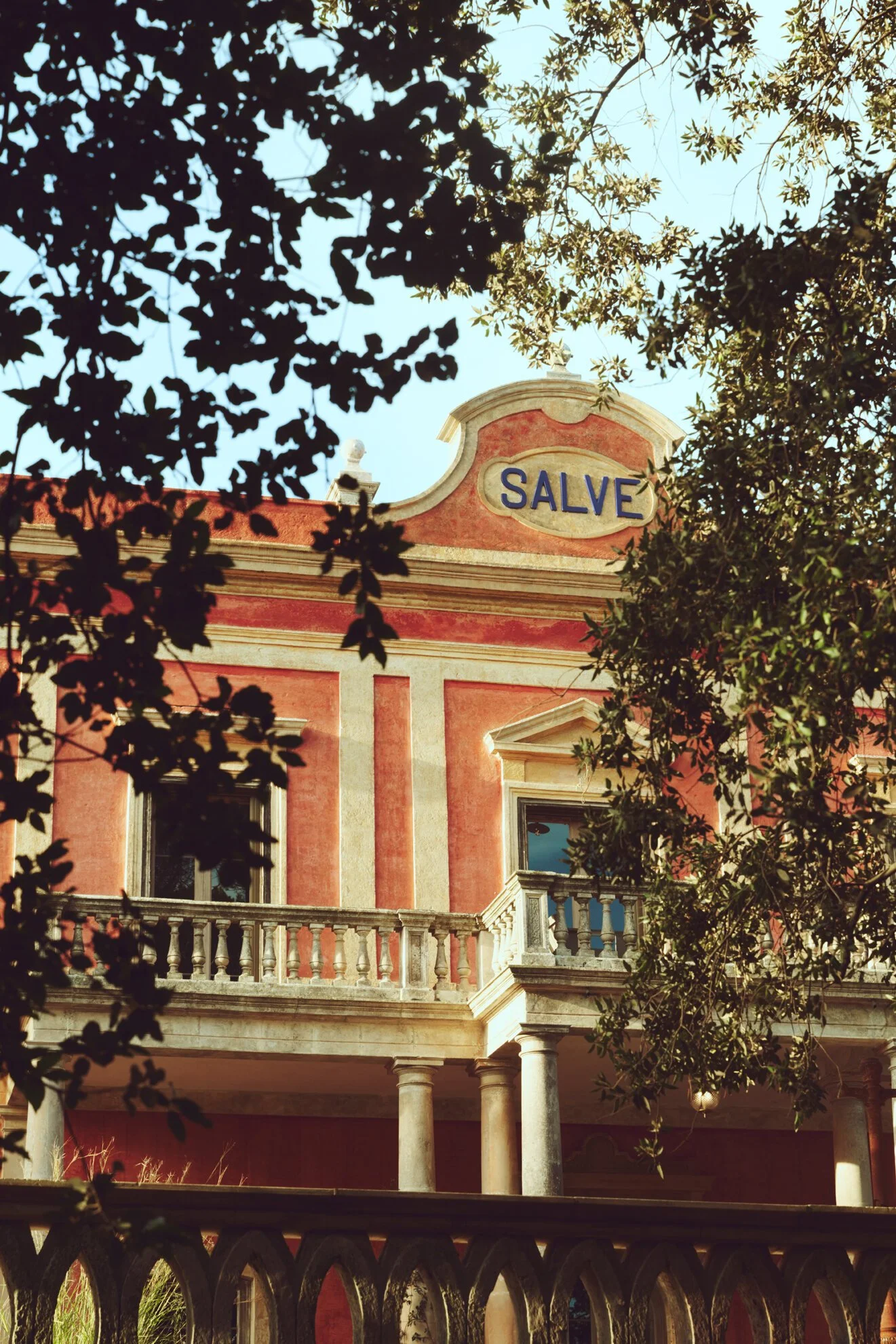A historic building with a pink facade, ornate balcony, and a sign that reads 'SALVE' at the top, partially obscured by tree foliage.