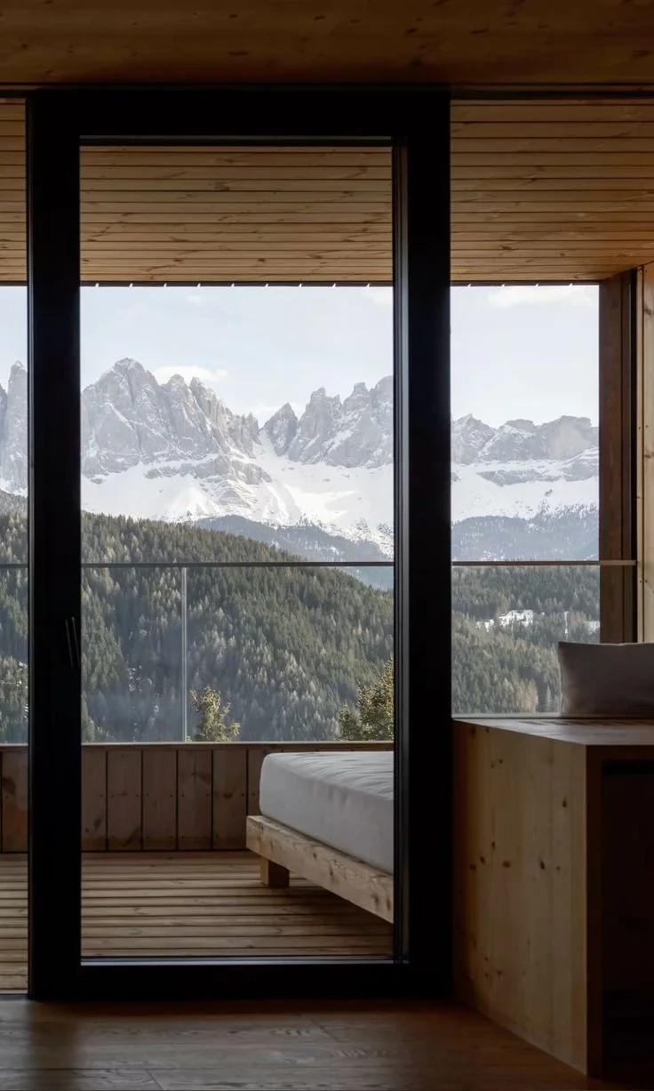 View of snow-capped mountains and green forest from a wooden room with a glass door and balcony.