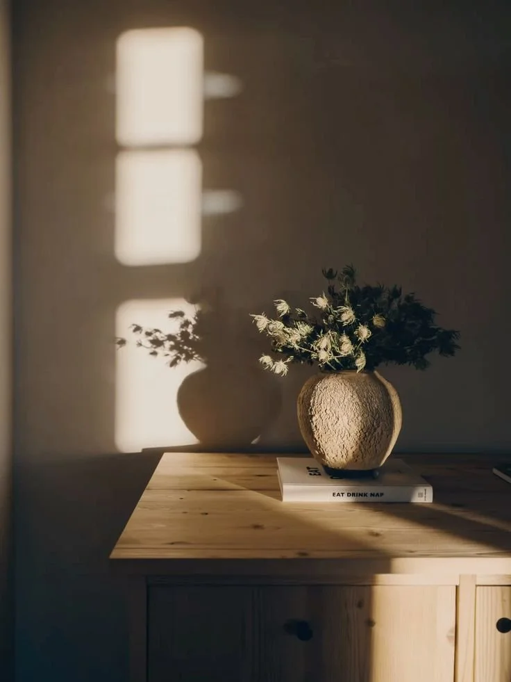Sunlight through window blinds casts a shadow on the wall, with a vase of flowers and a book on a wooden surface.