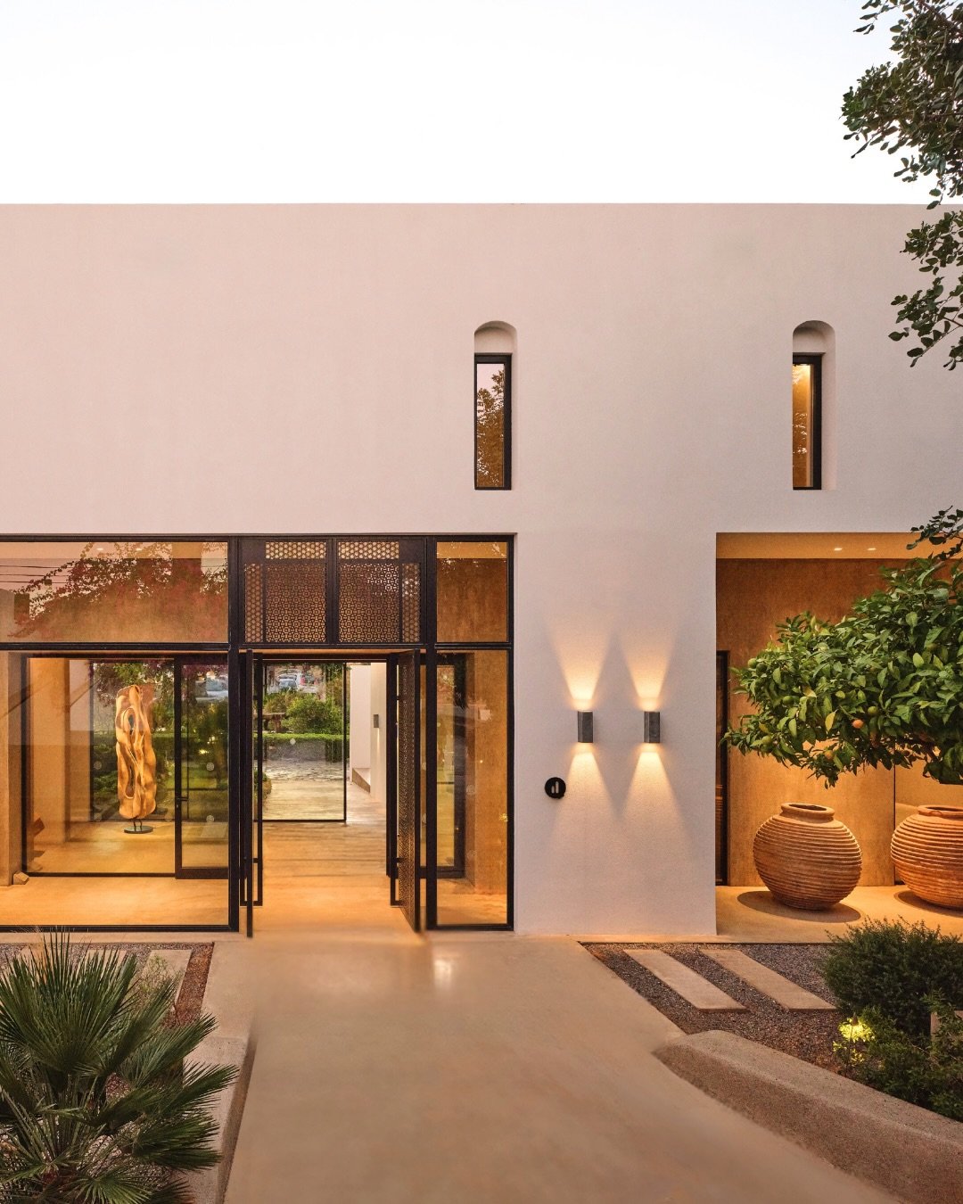 Modern house exterior at dusk with large glass doors, indoor art, and outdoor pottery, surrounded by minimal landscaping and trees.