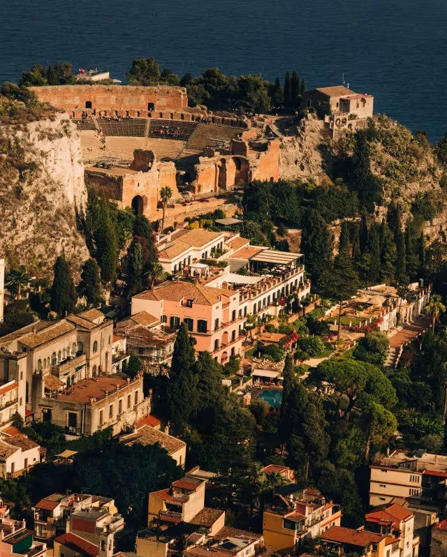 A hillside cityscape featuring historic ruins, terraced buildings, and lush greenery with the sea in the background.