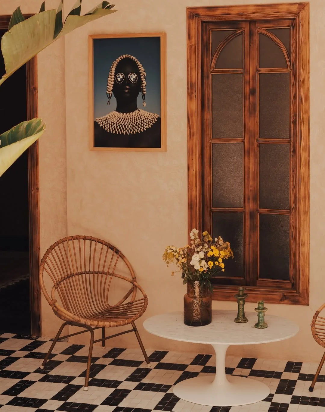 A cozy interior with a small white round table holding a vase of yellow and white flowers and two small green candles. Two rattan chairs are placed on a black and white checkered tile floor. On the wall, there's a colorful portrait of an African woman with beaded jewelry and an afro hairstyle, set inside a wooden frame. A wooden window with stained glass panels is behind the table.