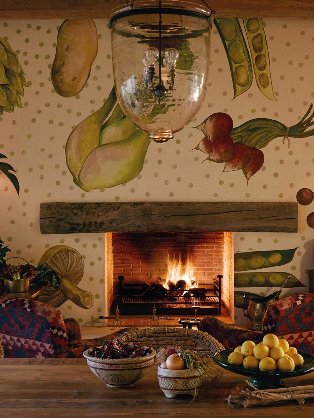 Living room with a brick fireplace and a fire burning, a wooden mantel, colorful patterned chairs, and a wall mural of vegetables including potatoes, carrots, onions, and peas. A glass chandelier hangs from the ceiling, and there are bowls of lemons, onions, and a basket on the wooden table in the foreground.