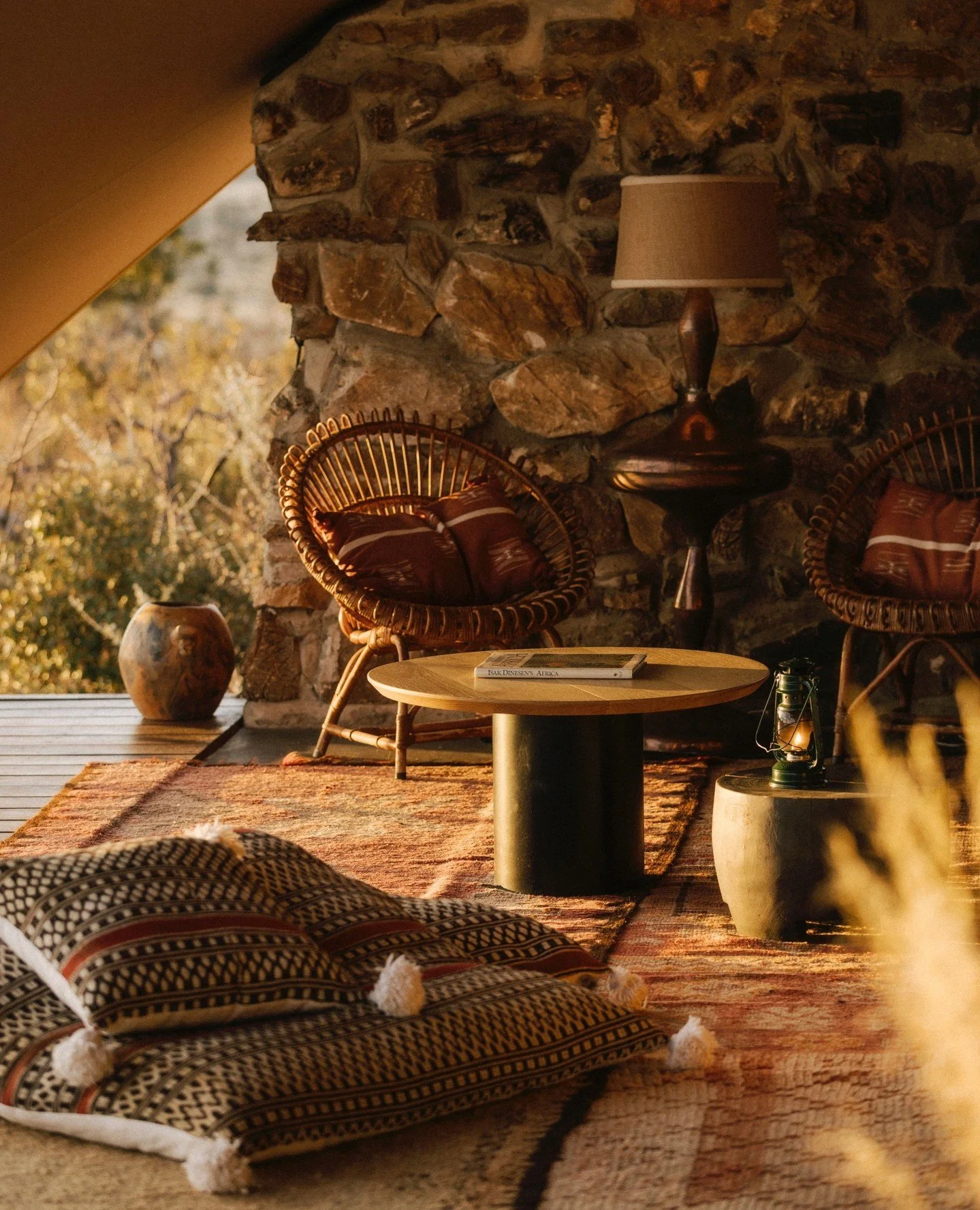 Cozy living room corner with two rattan chairs with cushions, a round coffee table, a stone wall, a lamp, a rug, and decorative vases, bathed in warm natural light.