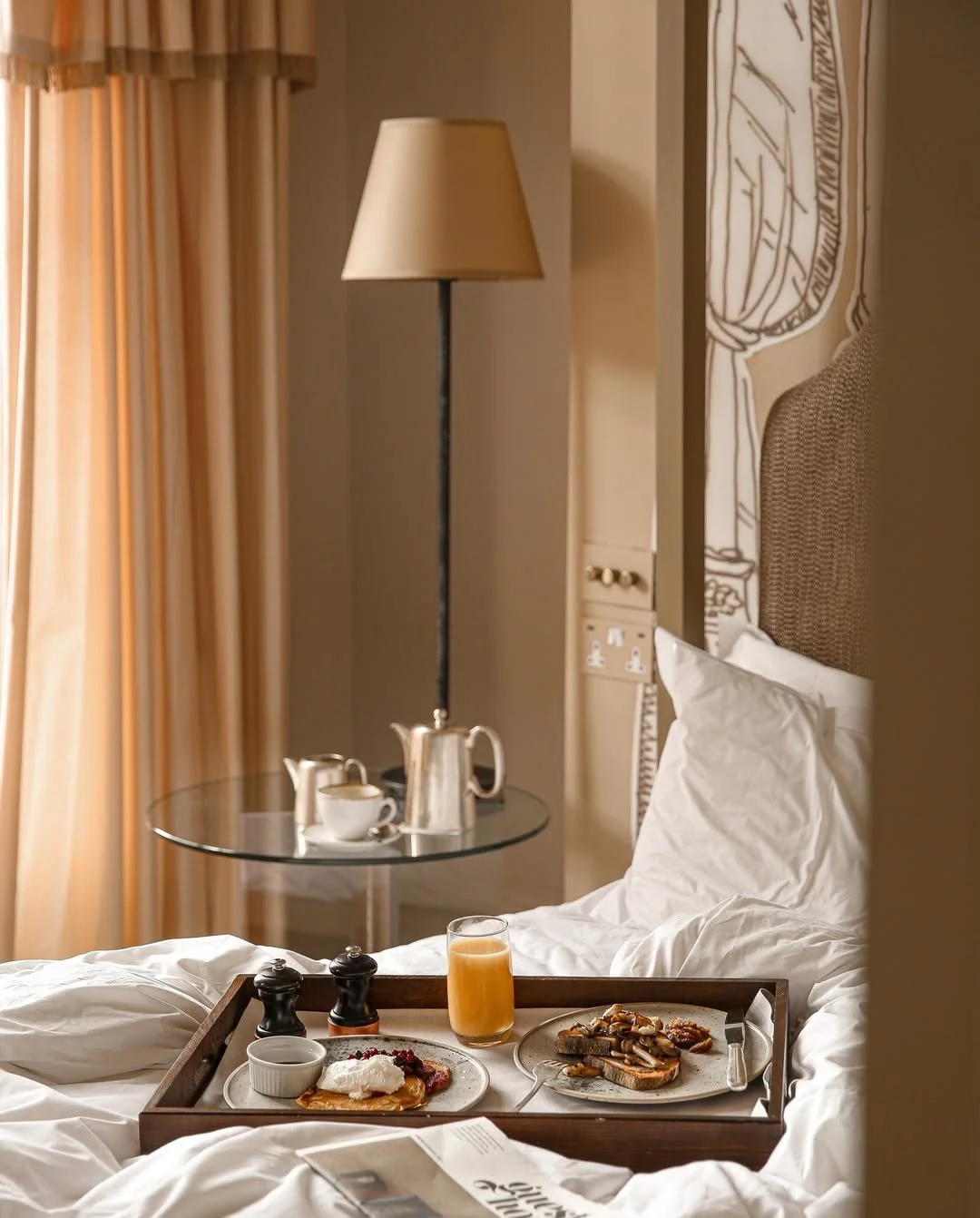 A hotel bed with a wooden breakfast tray containing a poached egg, fruit, a glass of orange juice, and a small dish of sauce or syrup. Next to the tray is a newspaper. In the background, a nightstand with a teapot, cups, and a lamp, and beige curtains.